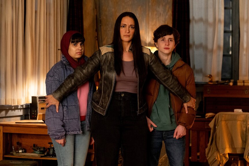 Asta Twelvetrees (Sara Tomko) stands and shields Sahar (Gracelyn Awad Rinke) and Max Hawthorne (Judah Prehn) on Resident Alien Episode 401.