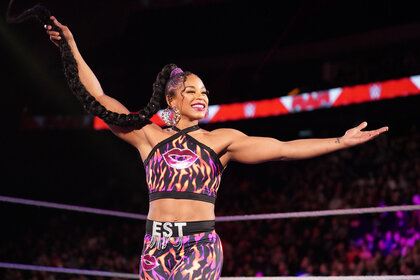 Bianca Belair holding her long braid in her right hand with both arms outstretched. She is smiling.