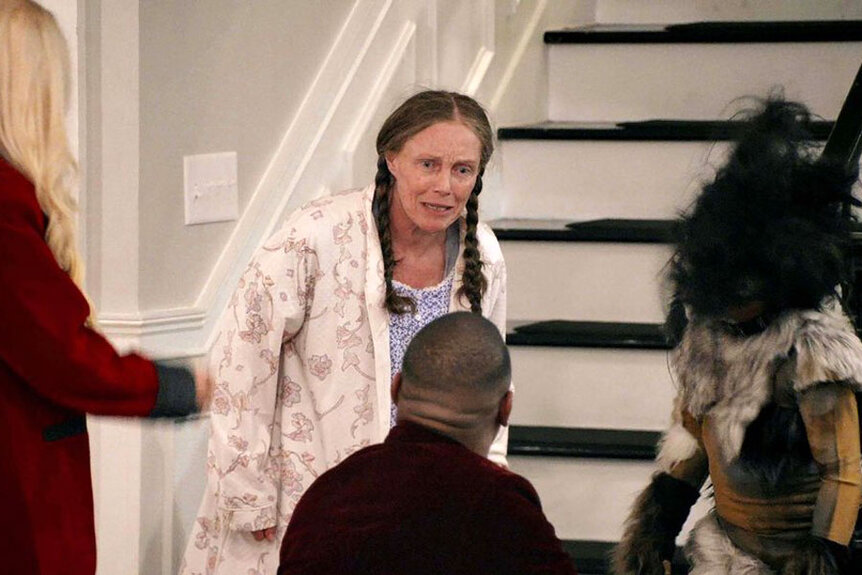 A woman in a robe wearing two braids looking upset as she talks to a man not facing the camera