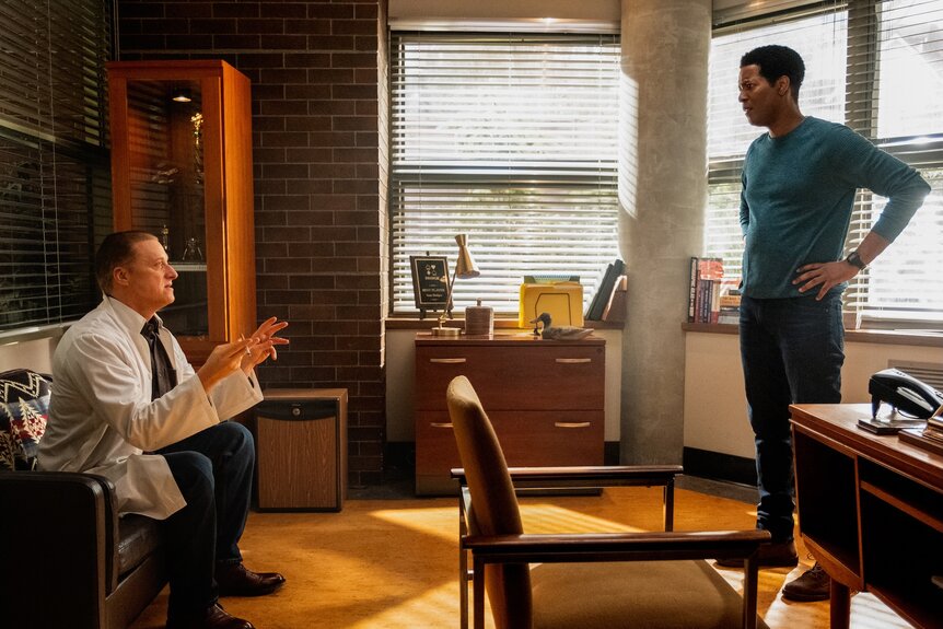 Harry Vanderspeigle (Alan Tudyk) sits on a couch and speaks with Sheriff Mike Thompson (Corey Reynolds) on Resident Alien Episode 401.