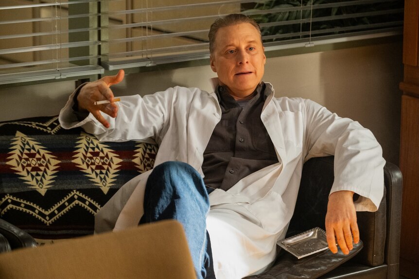 Harry Vanderspeigle (Alan Tudyk) speaks in his doctor's coat while reclining on a sofa on Resident Alien Episode 401.