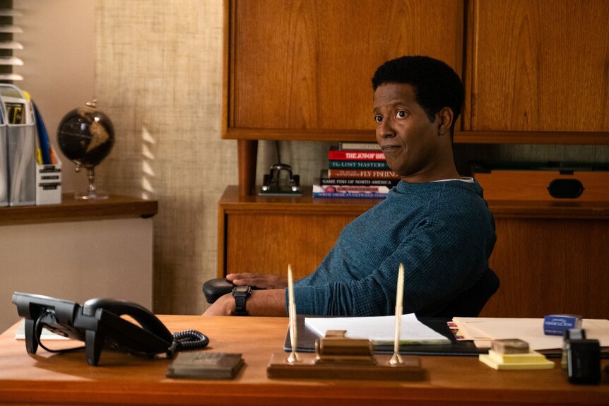 Sheriff Mike Thompson (Corey Reynolds) sits at a desk on Resident Alien Episode 401.