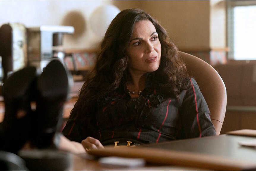 The Rainmaker's Lana Parilla as Bruiser Stone sat at desk with feet propped up